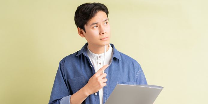 A thoughtful man holding a document and a pen, gazing upward, standing against a light yellow background.