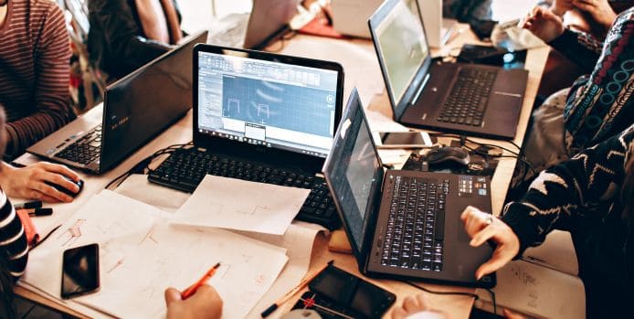 A group of people collaborating at a table with multiple laptops displaying design and technical software, surrounded by papers, phones, and pens.