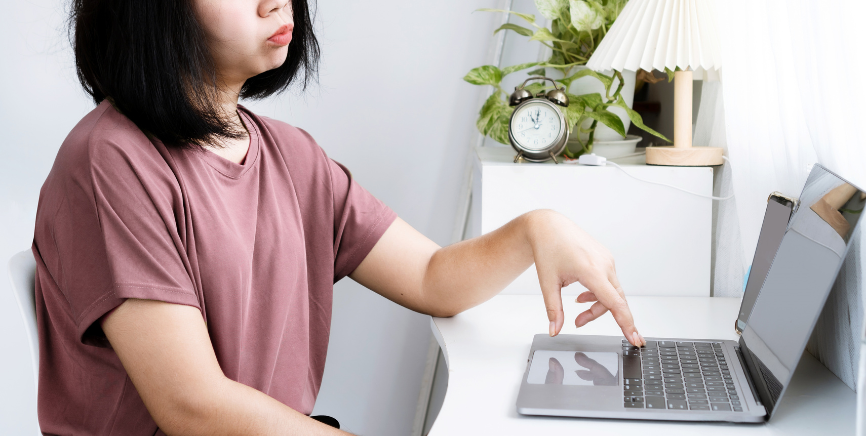 The image shows a woman sitting at a desk, pointing to her laptop with a frustrated expression. The setting includes a minimalistic workspace with a clock, a lamp, and a plant in the background, suggesting she is dealing with a technical issue like a frozen laptop.