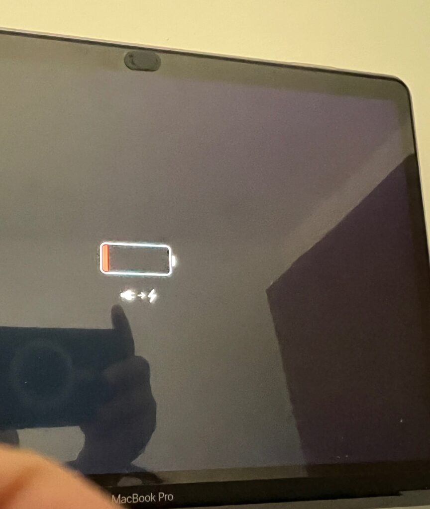 A MacBook not holding charge displaying a low battery warning with an almost empty battery icon and a charging symbol, indicating that the device needs to be plugged in. A reflection of a person holding a smartphone camera is visible on the screen.